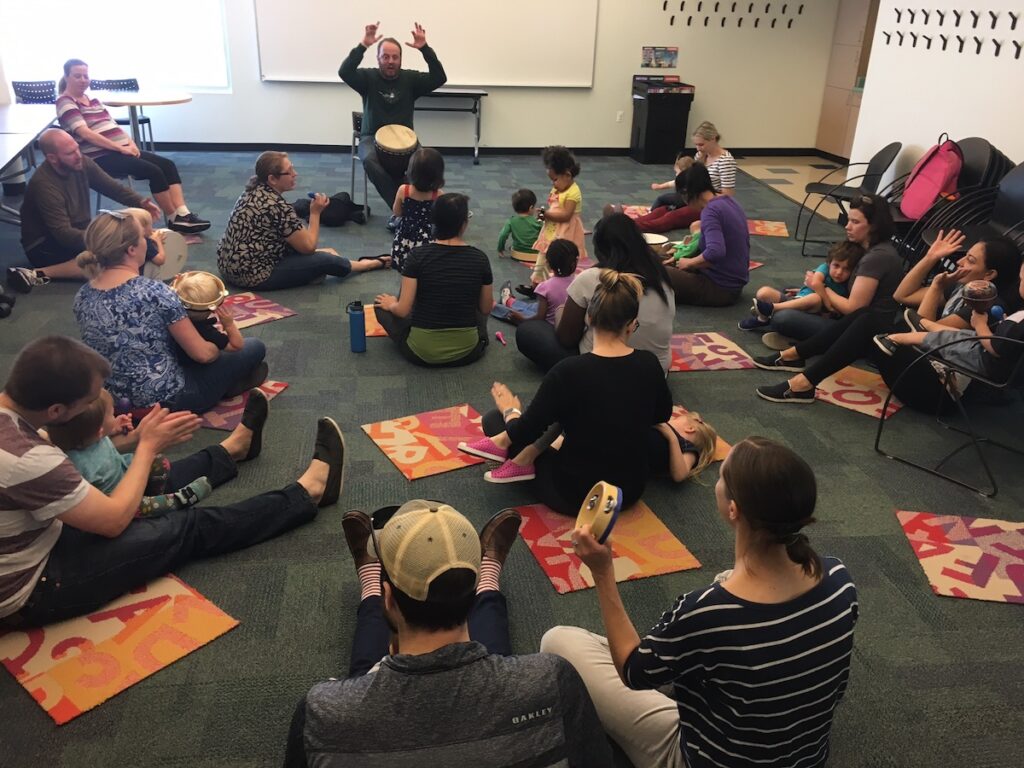 adults and children with site on the floor and clap along with man playing a drum