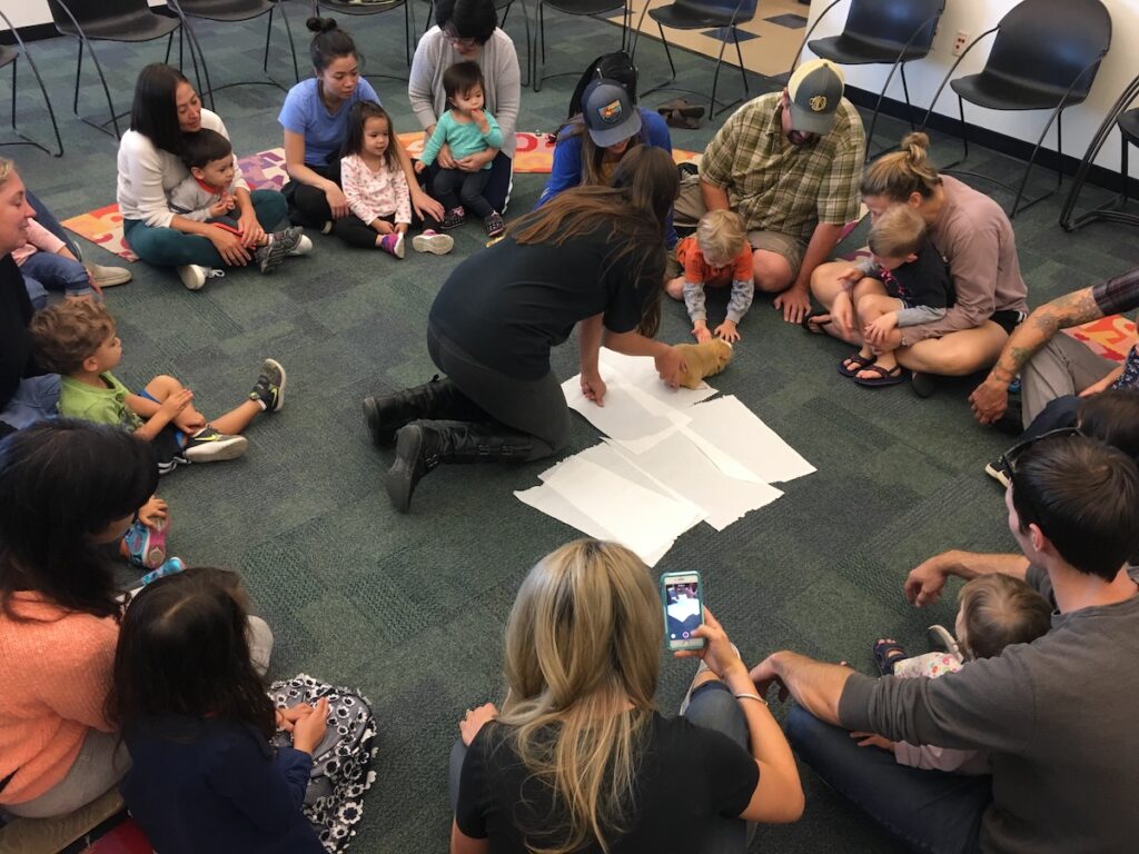 adults with children in their laps sit in a circle on the floor around a woman showing a child a small animal